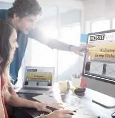 Two designers collaborate, pointing at a computer monitor that displays a website mockup next to a laptop