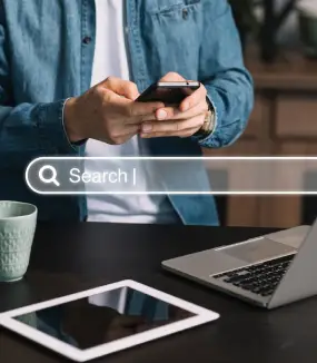 A person writes with a pen on a notepad while another works on a laptop, set against a brick wall background.